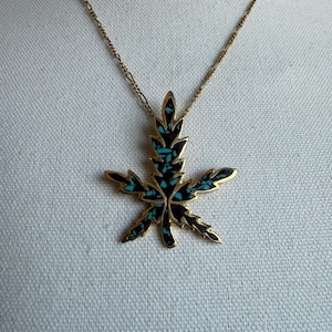 May include: Gold necklace with a cannabis leaf pendant. The pendant has a black and turquoise design, with a gold border. The chain is fine and gold. The necklace is shown against a white backdrop.