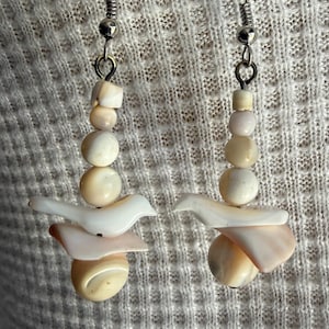 May include: A pair of drop earrings featuring silver-toned metal hooks and a vertical arrangement of cream and pale peach-colored shell beads. Each earring includes a small bird-shaped shell carving, flat shell pieces, and round beads.