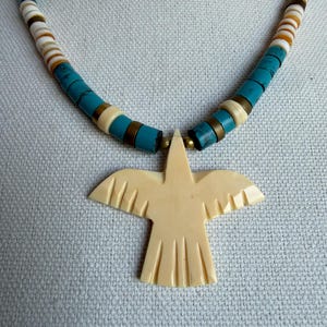 Puede incluir: Un collar de cuentas turquesas con un colgante de pájaro color crema. El collar presenta cuentas turquesas, blancas y marrones alternas. El colgante de pájaro está tallado con detalles de alas y cola, sobre fondo azul claro.