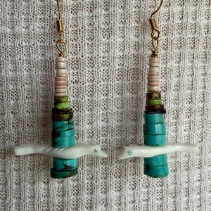 May include: A pair of dangle earrings featuring gold-tone hooks and a vertical stack of beads. The beads include light pink, green, dark brown, and turquoise discs, accented by a horizontal white bird-shaped bead with small red and blue details.