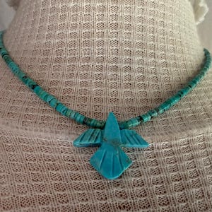 May include: A turquoise necklace featuring a series of small, flat, circular beads strung together, centered with a carved turquoise pendant shaped like a bird with outstretched wings and detailed linear etchings on its surface.