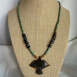 May include: A necklace featuring a dark brown carved bird pendant suspended from a strand of small green beads. The necklace includes segments of dark brown disc-shaped beads and two light tan cylindrical beads near the pendant attachment point.