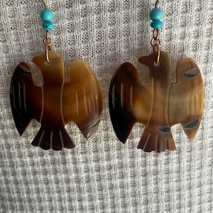 Puede incluir: Un par de pendientes colgantes con colgantes de cuerno marrón tallado en forma de aves con las alas extendidas. Cada pendiente cuelga de un gancho de color cobre y está decorado con dos pequeñas cuentas redondas de color azul turquesa.