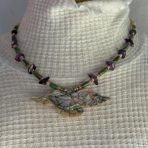 Könnte beinhalten: Eine Halskette mit einem zentralen Anhänger in Vogelform aus irisierendem Abalone-Muschelmaterial. Die Kette besteht aus abwechselnden grünen zylindrischen Perlen, kleinen braunen Zwischenperlen und unregelmäßigen violetten Amethystsplittern.