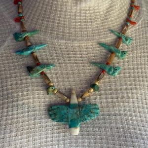 May include: A beaded necklace featuring a central turquoise bird pendant with white stone accents. The strand consists of carved turquoise bird-shaped beads interspersed with small cylindrical tan beads, bright orange coral-like segments, and tiny turquoise spheres.