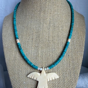 Puede incluir: Un collar de cuentas turquesas con un colgante de pájaro tallado en color marfil. El collar se exhibe en un maniquí marrón. El colgante de pájaro tiene alas extendidas y tallas de plumas detalladas. Es una joya única.