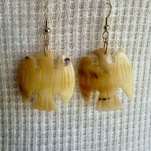 May include: A pair of drop earrings featuring carved, cream-coloured bird-shaped pendants with brown marbled accents. The pendants are suspended from gold-toned metal fishhook wires, showcasing detailed wing and tail feather engravings.