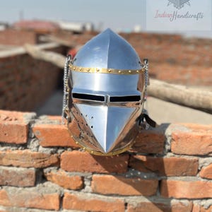 May include: A polished silver knight's helmet with a pointed top and a hinged visor. The helmet features brass accents and chainmail details. The image is set against a brick wall and includes the text "Indian Handicrafts".