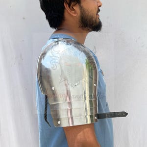 Pode incluir: Uma ombreira de metal prateado polido, parte de um conjunto de armadura medieval. A ombreira é feita de placas sobrepostas e é presa a uma tira preta. A armadura é usada no braço de uma pessoa.