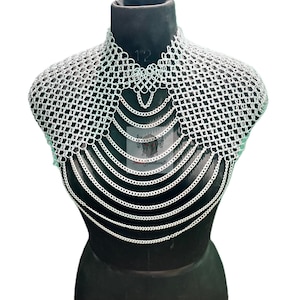 Puede incluir: Un collar de cota de malla plateada exhibido en un maniquí negro. El collar presenta un diseño de malla tejida alrededor del cuello y los hombros, con múltiples hilos de cadenas que caen en cascada sobre el pecho. El metal tiene un acabado brillante y reflectante.
