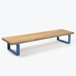 May include: A rectangular wooden bench with a natural wood grain finish. The bench is supported by two blue, U-shaped metal legs. The design is minimalist and modern, suitable for various interior settings.