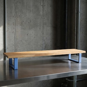 May include: A rectangular wooden shelf with a light brown finish. The shelf is supported by two light blue metal frames. The shelf is placed on a stainless steel surface, with a concrete wall in the background.
