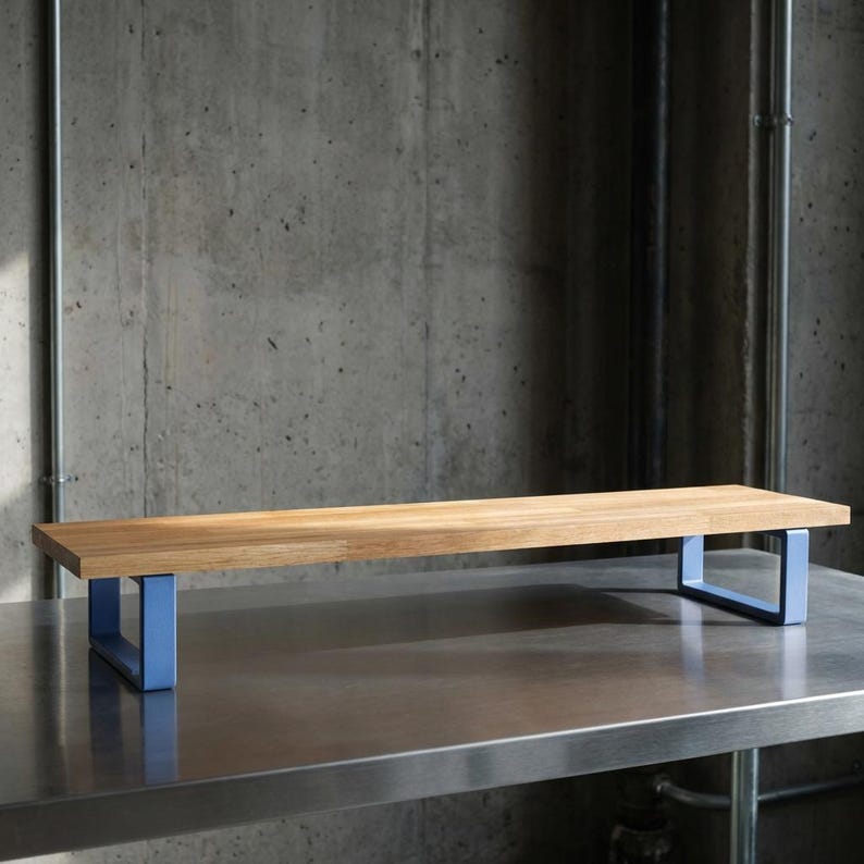 May include: A rectangular wooden shelf with a light brown finish. The shelf is supported by two light blue metal frames. The shelf is placed on a stainless steel surface, with a concrete wall in the background.