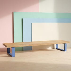 May include: A wooden bench with a light brown top and blue metal legs. The bench is rectangular and sits on a light-coloured wooden floor. The background features pastel-coloured rectangular panels in pink, green, blue, and cream.