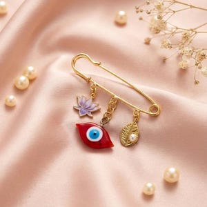 May include: A gold-colored safety pin with dangling charms. Charms include a red evil eye, a purple lotus flower, and a gold leaf with a pearl. The pin is on a pink satin surface with scattered pearls.