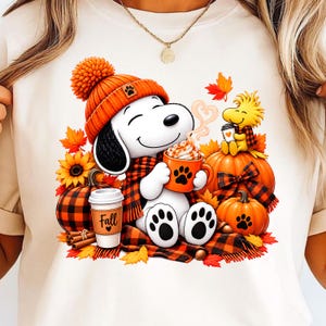 Może przedstawiać: Kremowy t-shirt z grafiką przedstawiającą Snoopy'ego w pomarańczowej czapce i szaliku, trzymającego pomarańczowy kubek z odciskiem łapy. Woodstock siedzi na dyni. Wzór zawiera jesienne liście, słoneczniki oraz kubek z napisem Fall.