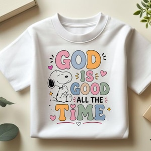 Puede incluir: Camiseta blanca con Snoopy de Peanuts y la frase "GOD IS GOOD ALL THE TIME" en letras de colores. El dise&ntilde;o incluye corazones y estrellas, creando un aspecto alegre e inspirador.