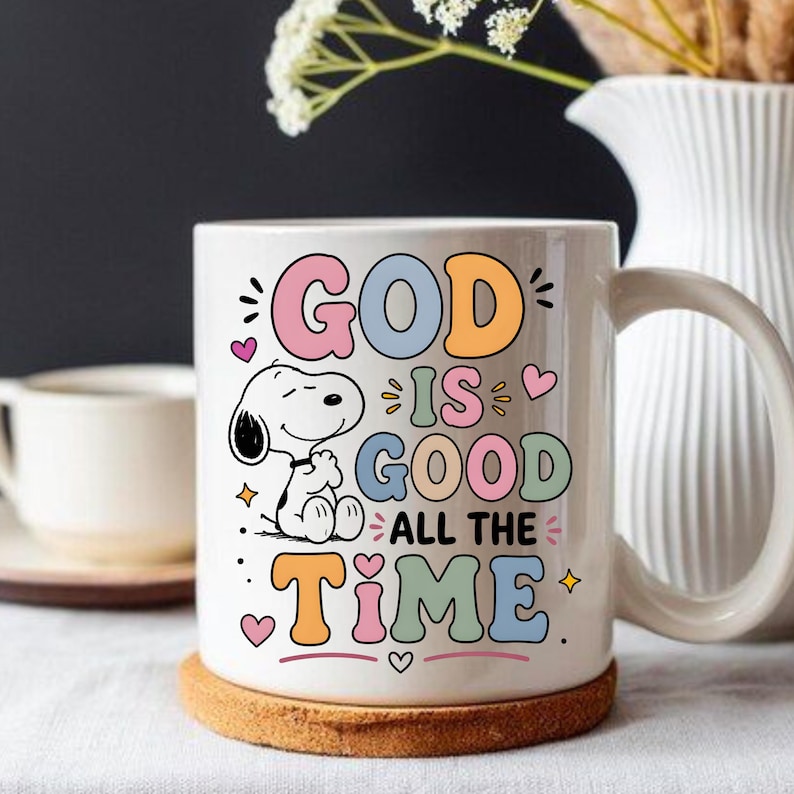 Puede incluir: Taza de cer&aacute;mica blanca con la frase "GOD IS GOOD ALL THE TIME" en letras de colores. Tambi&eacute;n aparece un personaje de dibujos animados de Snoopy en la taza. La taza est&aacute; sobre un posavasos redondo de corcho.