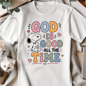 Puede incluir: Camiseta blanca con la frase "GOD IS GOOD ALL THE TIME" en letras de colores. Un personaje de dibujos animados de Snoopy est&aacute; incluido en el dise&ntilde;o. La camiseta tiene cuello redondo y mangas cortas.