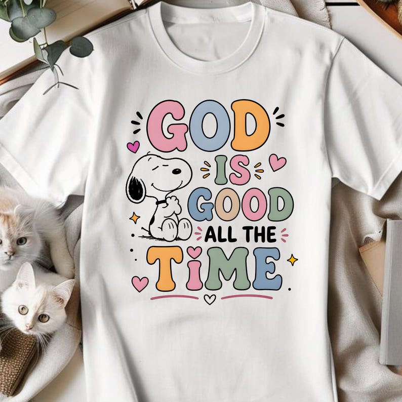 Puede incluir: Camiseta blanca con la frase "GOD IS GOOD ALL THE TIME" en letras de colores. Un personaje de dibujos animados de Snoopy est&aacute; incluido en el dise&ntilde;o. La camiseta tiene cuello redondo y mangas cortas.