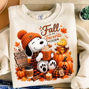 May include: A cream-colored sweatshirt featuring a graphic of Snoopy wearing an orange beanie, holding an orange mug labeled 'COZY DAYS AHEAD'. Text reads 'Fall IS MY Favorite COLOR', 'SWEATER WEATHER COFFEE & LEAVES', 'BUT FIRST COFFEE', and 'STAY COZY'.