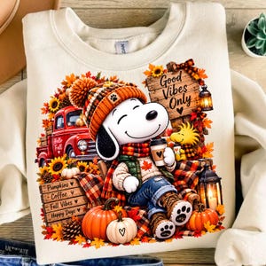 May include: A cream-coloured sweatshirt featuring a graphic of a cartoon dog wearing a knit hat and scarf, holding a coffee cup. The design includes a red truck, pumpkins, sunflowers, lanterns, and wooden signs with the text Good Vibes Only, Pumpkins, Coffee, Fall Vibes, and Happy Days.