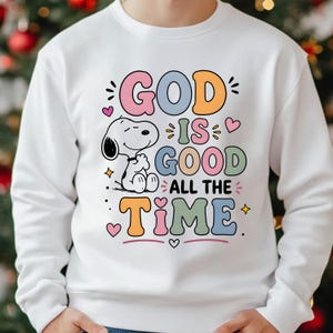 Puede incluir: Sudadera blanca con la frase "GOD IS GOOD ALL THE TIME" en letras de colores. Un personaje de dibujos animados de Snoopy aparece a la izquierda, con corazones y estrellas. La sudadera es de cuello redondo.