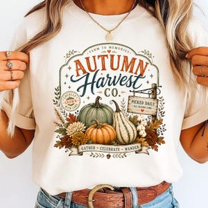 Pode incluir: Uma t-shirt de cor creme com uma impressão gráfica com o texto Autumn Harvest Co. Farm to Memories. O design inclui ilustrações de abóboras, cabaças, folhas de outono, flores e uma placa de madeira com a inscrição Picked Daily All Season Long.