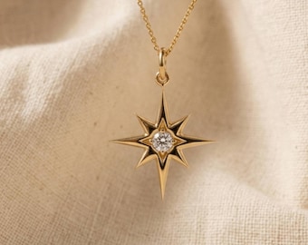 May include: A gold-colored necklace with a starburst pendant. The pendant features a central, round, clear gemstone and is suspended from a delicate chain. The starburst design has pointed edges and a dark outline, set against a neutral fabric background.