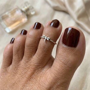 May include: A silver toe ring with a dragonfly design. The ring is worn on a toe with dark brown nail polish. The image is a close-up shot, highlighting the ring's details and the nail polish color.