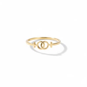 May include: A gold-colored ring featuring a double female symbol design. The ring has a thin band and the symbols are connected in the center. The ring is set against a white background.