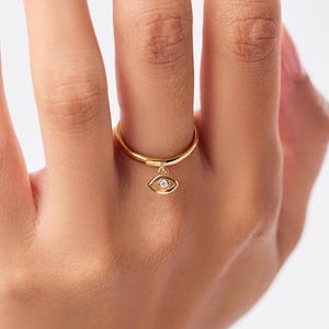 May include: A gold-colored ring with a small eye-shaped charm. The charm has a clear stone in the center. The ring is on a finger.