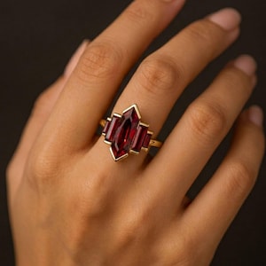 Peut inclure: Bague en or ornée d'une grande pierre rouge foncé rectangulaire au centre, flanquée de pierres rouges rectangulaires plus petites. La bague a un design géométrique, sur fond sombre.
