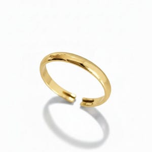 Puede incluir: Un anillo abierto de color dorado. El anillo tiene una banda lisa y redondeada con un pequeño hueco en la parte delantera. El anillo está sobre un fondo blanco, con una sombra proyectada debajo.