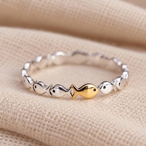 Op de afbeelding: Een smalle ring met een herhalend patroon van kleine, gestileerde visvormen. Het merendeel van de vissen is zilverkleurig, terwijl één enkele vis nabij het midden van de ring een contrasterende, gepolijste gouden afwerking heeft.