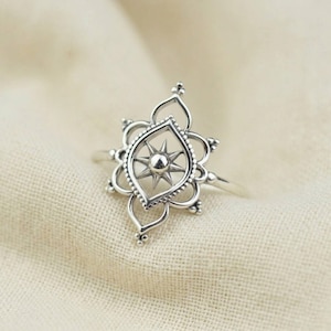 May include: A silver ring featuring an openwork design with a central starburst motif. The ring's focal point is a stylized star within a teardrop-shaped frame, surrounded by a floral-inspired border. The ring is set against a neutral fabric background.