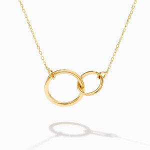 Può includere: Una collana dorata con due cerchi intrecciati. I cerchi sono collegati e sospesi a una delicata catena. La collana è mostrata su uno sfondo bianco, con una sottile ombra.