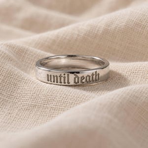 May include: A polished silver-toned metal band ring featuring the engraved text "until death" in a gothic-style font. The ring has a smooth, reflective surface and a flat profile, showcasing clear, dark lettering etched into the outer circumference.
