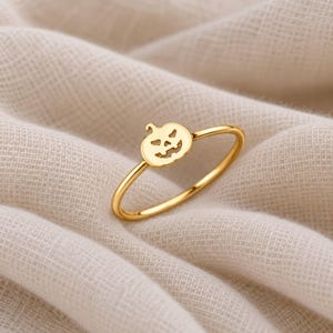 May include: A thin gold ring featuring a small, flat, cutout pumpkin charm with a carved jack-o'-lantern face. The band is smooth and circular, showcasing a polished metallic finish that reflects light across its surface.
