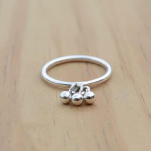 Puede incluir: Un anillo de plata con tres pequeñas esferas colgantes. El anillo es fino y circular, con las esferas agrupadas en la parte inferior. El anillo está sobre una superficie de madera marrón claro.