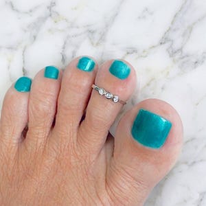 May include: A silver toe ring with three clear, round gemstones is worn on a foot with teal nail polish. The foot is positioned against a white and gray marble background. The ring is delicate and minimalist.