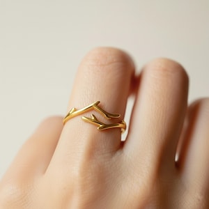 Könnte beinhalten: Ein goldfarbener Ring mit einem zweigartigen Design. Der Ring umschließt den Finger, wobei sich zwei Zweige nach außen erstrecken. Der Ring besteht aus einem glänzenden, metallischen Material. Der Ring ist ein Schmuckstück.