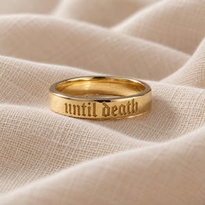 May include: A gold-colored metal band ring featuring the engraved text "until death" in a gothic-style font. The ring has a smooth, polished finish and a flat outer surface, showcasing the lettering clearly across the front of the band.