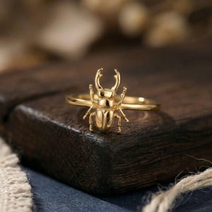 May include: A gold-colored ring featuring a detailed beetle design. The ring's band is thin, and the beetle has prominent antennae and legs. The ring is displayed on a dark wooden surface, showcasing the intricate details of the jewelry.