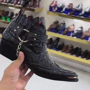 Peut inclure: Une bottine noire en cuir avec une texture peau de crocodile. La botte présente un bout pointu avec un accent métallique, une sangle avec des détails argentés et une petite chaîne. La botte est tenue devant un magasin de chaussures.