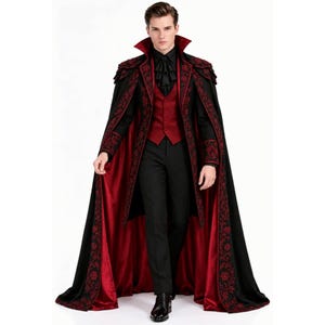 Puede incluir: Una capa negra hasta el suelo con forro de satén rojo y un intrincado bordado floral rojo en los bordes. El conjunto incluye un chaleco rojo oscuro, una camisa negra con cuello de volantes y pantalones negros, creando un atuendo formal.