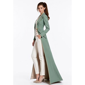 May include: A sage green floor-length coat with a tailored fit, long sleeves, and a notched lapel. The coat features a single-breasted front with gold-toned buttons and a contrasting beige lining. It is worn over a white top and white trousers.