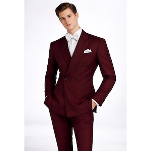 May include: A burgundy double-breasted suit with a white dress shirt, bow tie, and pocket square. The suit has a tailored fit and is worn with the hands in the pockets. The background is white.