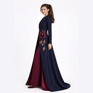May include: A floor-length navy blue coat with long sleeves and a single button closure, featuring an embroidered burgundy flower with gold leaves on the front. The coat is worn over a solid burgundy floor-length dress with a smooth finish.
