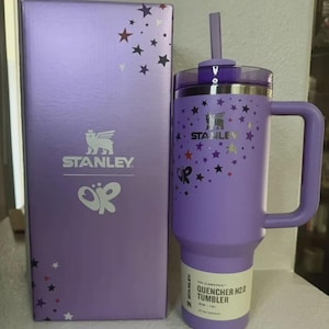 K&ouml;nnte beinhalten: Ein lila Stanley Quencher H2.0 Becher mit Griff und Strohhalm, neben einer passenden Box. Der Becher und die Box weisen ein Sternmuster und das Stanley-Logo auf. Der Becher fasst 1,18L.